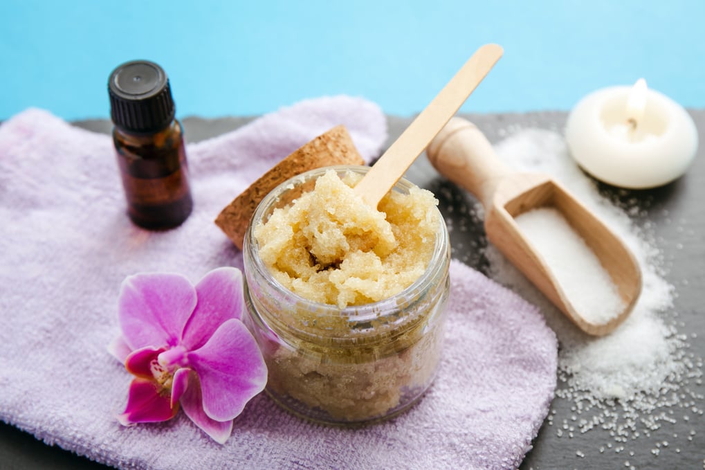 Side view of homemade sugar scrub in glass jar with ingredients for decoration, sugar on spoon and aroma oil, essential oil on blue background. Pink orchid blossom for decoration.